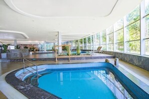 An indoor swimming pool with clear blue water, surrounded by a tiled deck and seating area, with large windows providing natural light. - Qingdao Tiantai Mountain le'ai Hot Spring Art Hotel (Qingdao)