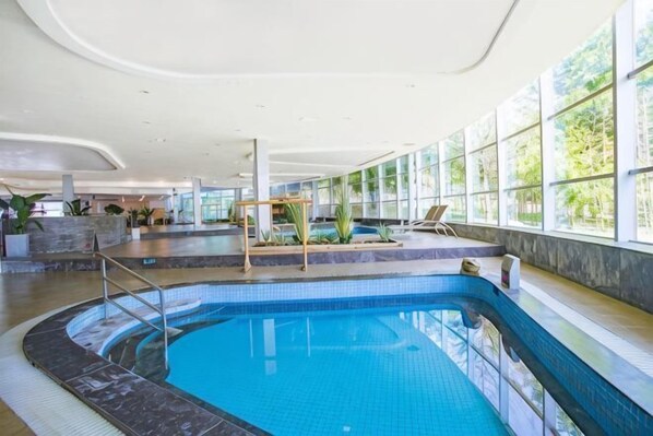 An indoor swimming pool with clear blue water, surrounded by a tiled deck and seating area, with large windows providing natural light. - Qingdao Tiantai Mountain le'ai Hot Spring Art Hotel (Qingdao)