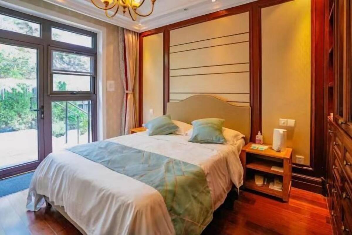 A bedroom with a bed, bedside tables, a nightstand, a window, and a view of greenery.