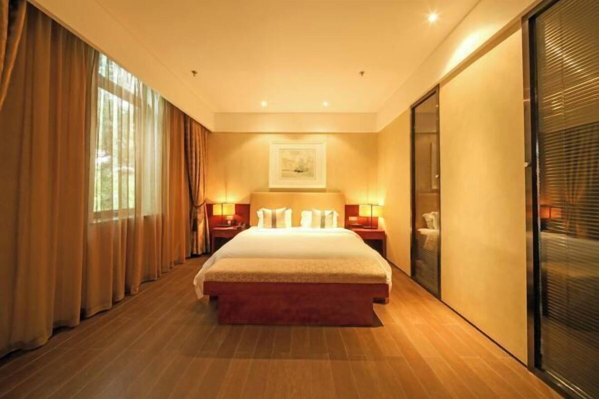 A hotel room with a large bed, two bedside tables with lamps, a large window with curtains, and a sliding glass door.