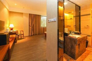 A hotel room with a wooden floor, a desk with a lamp, a chair, a large mirror, and a bathroom with a marble sink and shower. - Qingdao Tiantai Mountain le'ai Hot Spring Art Hotel (Qingdao)