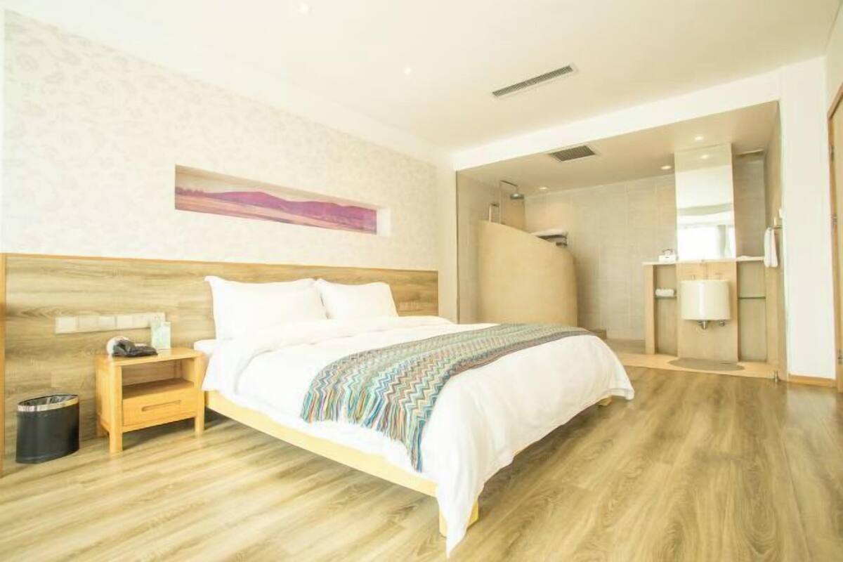 A modern hotel room with a wooden bed, a nightstand, a trash bin, a bathroom with a mirror and a towel rack.