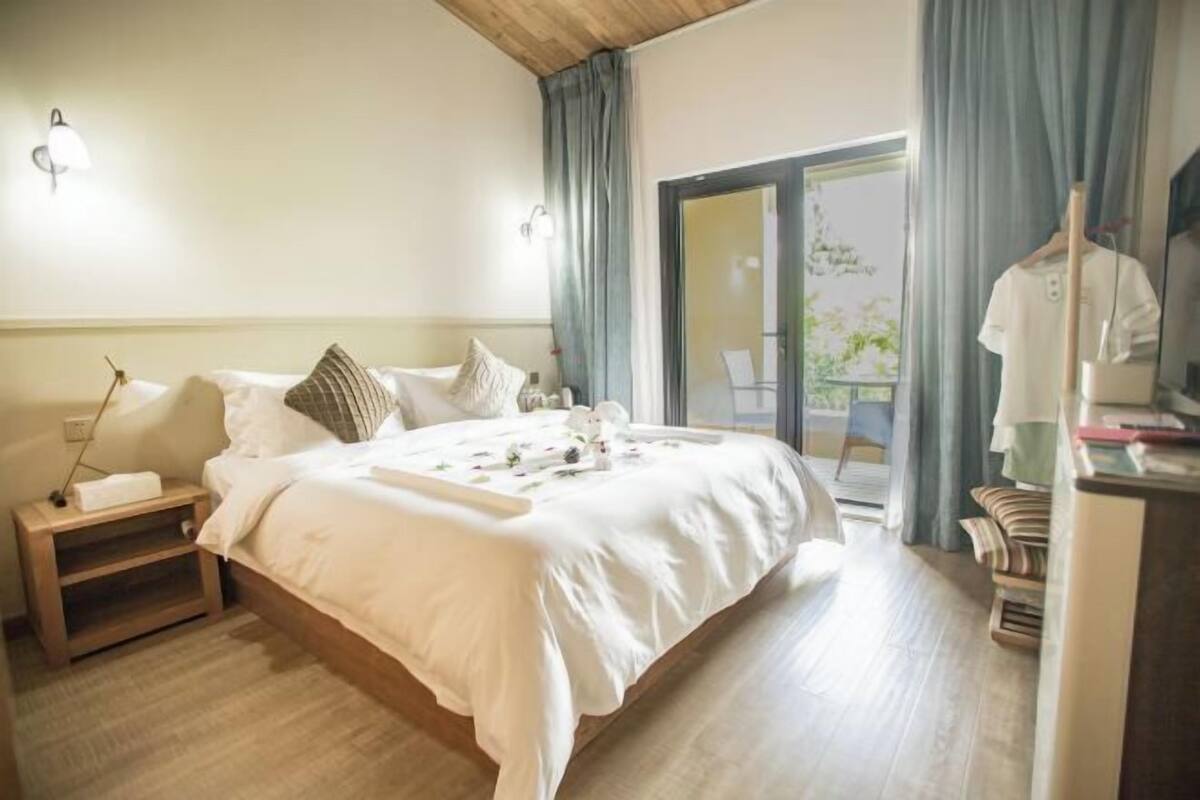 A bedroom with a bed, bedside table, lamp, and a view of the outside.
