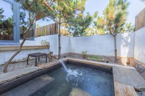 A small outdoor hot tub with a wooden bench and a tree in the background.