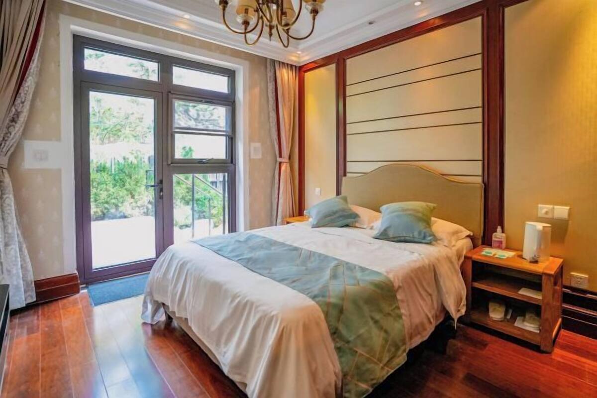 A bedroom with a large bed, a nightstand, a chandelier, and a view through a glass door.