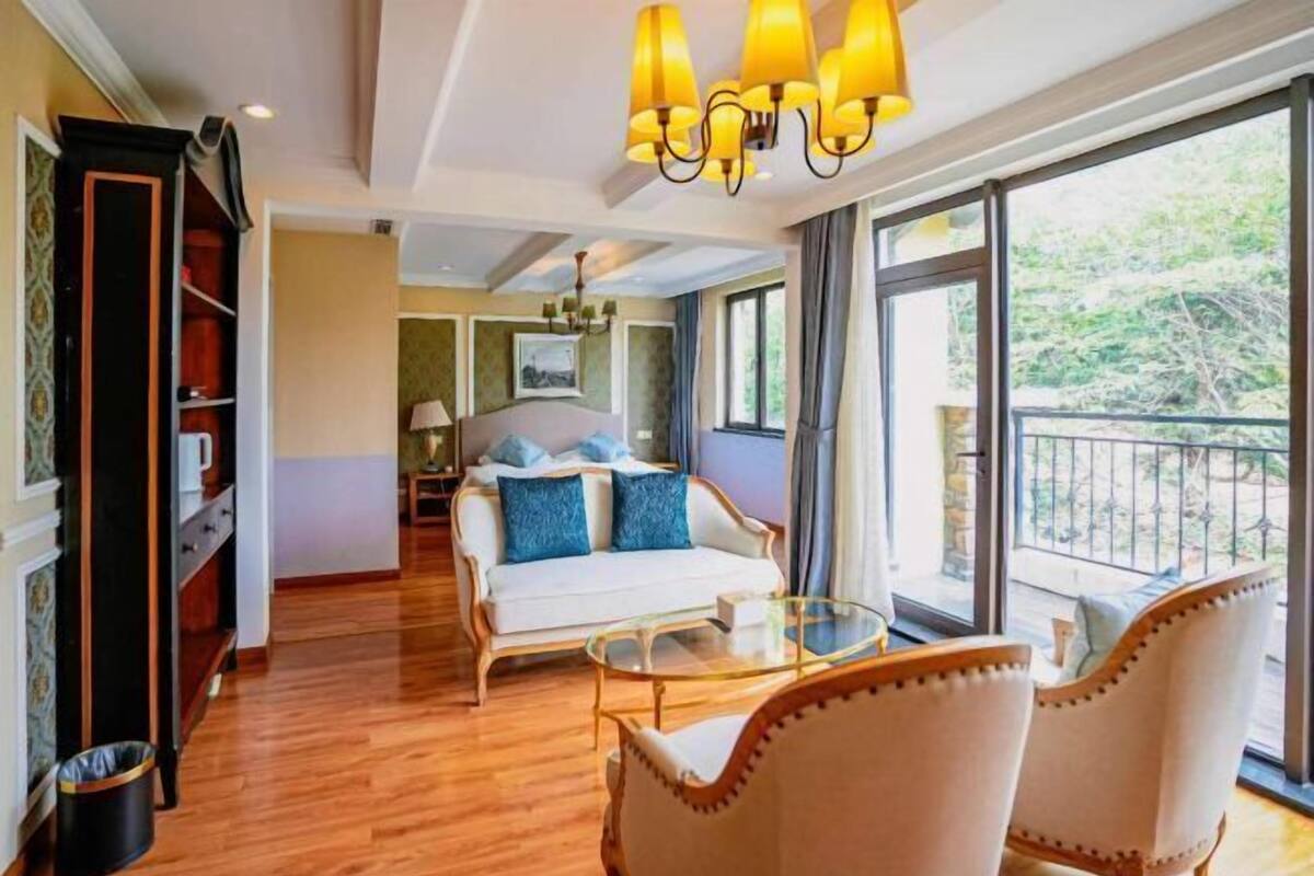 A hotel room with a wooden floor, a white sofa with blue cushions, a glass coffee table, and a balcony with a railing.