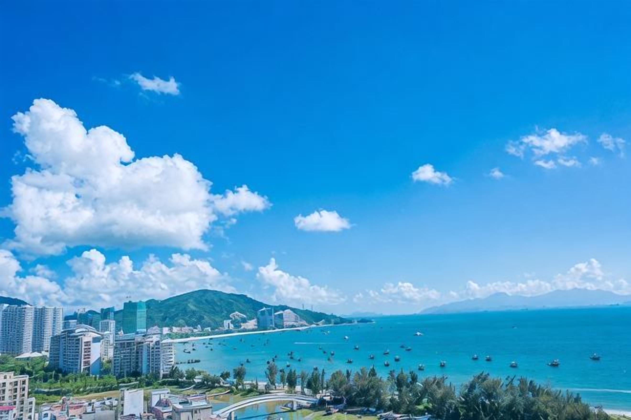A coastal cityscape with buildings, a clear blue sky, and a calm sea with boats.