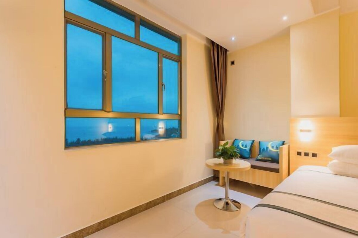A hotel room with a large window, a small round table, a sofa with cushions, and a bed.