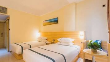 Quarto de hotel com duas camas, cabeceira de madeira, uma mesa redonda pequena com uma planta e um quadro na parede.