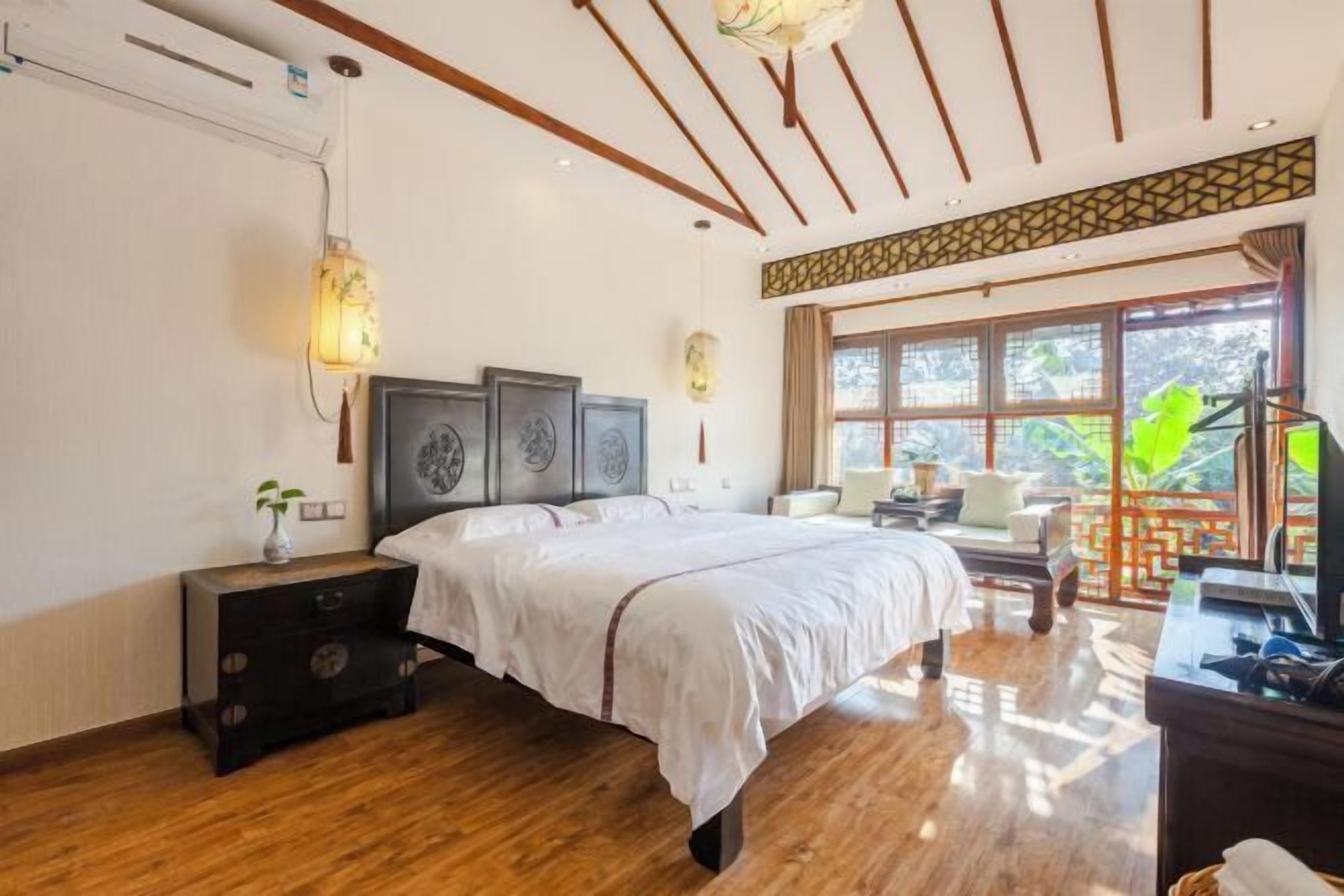 A bedroom with a wooden floor, a bed with a white bedspread, a nightstand with a vase, and a window with a view of greenery.