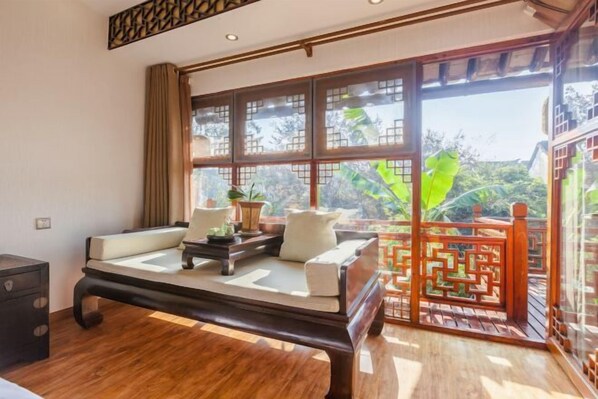 A room with a wooden bed, a small table with a lamp, and a large window with a view of greenery. - Pushe Guiyin Inn (Kunshan)