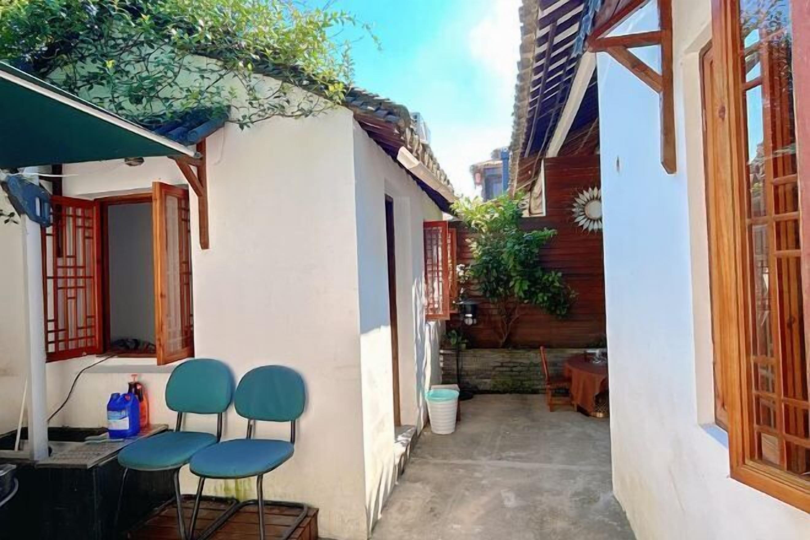 A narrow alleyway with a white building on the left and a wooden building on the right. There are two blue chairs outside the white building, and a small table with a red tablecloth in the alley.