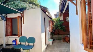 A narrow alleyway with a white building on the left and a wooden building on the right. There are two blue chairs outside the white building, and a small table with a red tablecloth in the alley.