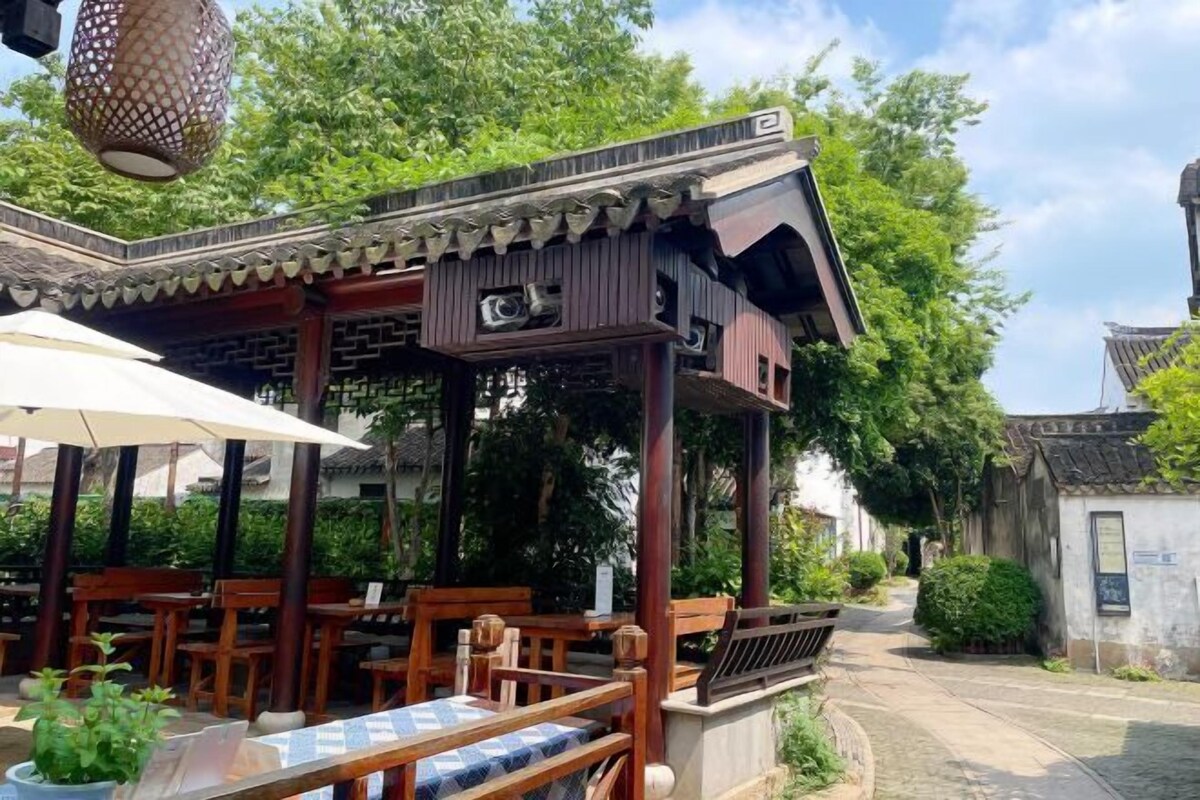A traditional wooden pavilion with a tiled roof, surrounded by greenery and a paved path.