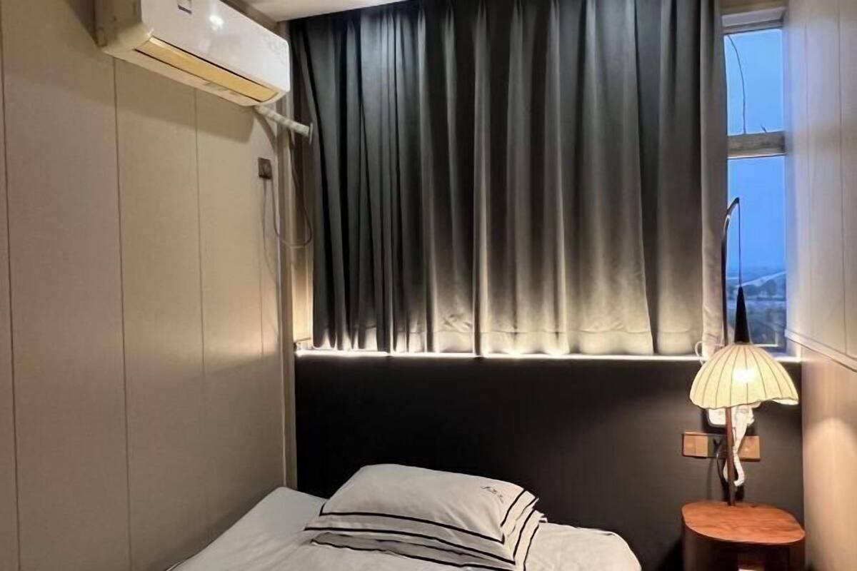 Hotelkamer met een bed, een raam met gordijnen, een lamp op een nachtkastje en een airconditioning op de muur.