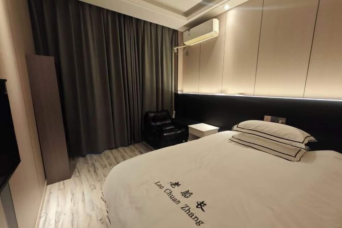 Een hotelkamer met een bed, een stoel, een klein tafeltje en een luchtverkoeler aan de muur.