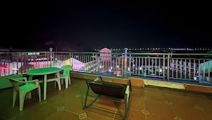 A rooftop terrace with a view of a cityscape at night, featuring a railing, a table, and chairs.