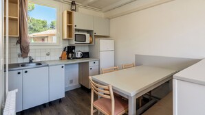 A compact kitchen with a microwave, coffee maker, and a small refrigerator. There is a dining table with chairs and a window with a view of a building outside. - Cottage with pool in Provence N°2 (La Roque-sur-Pernes)