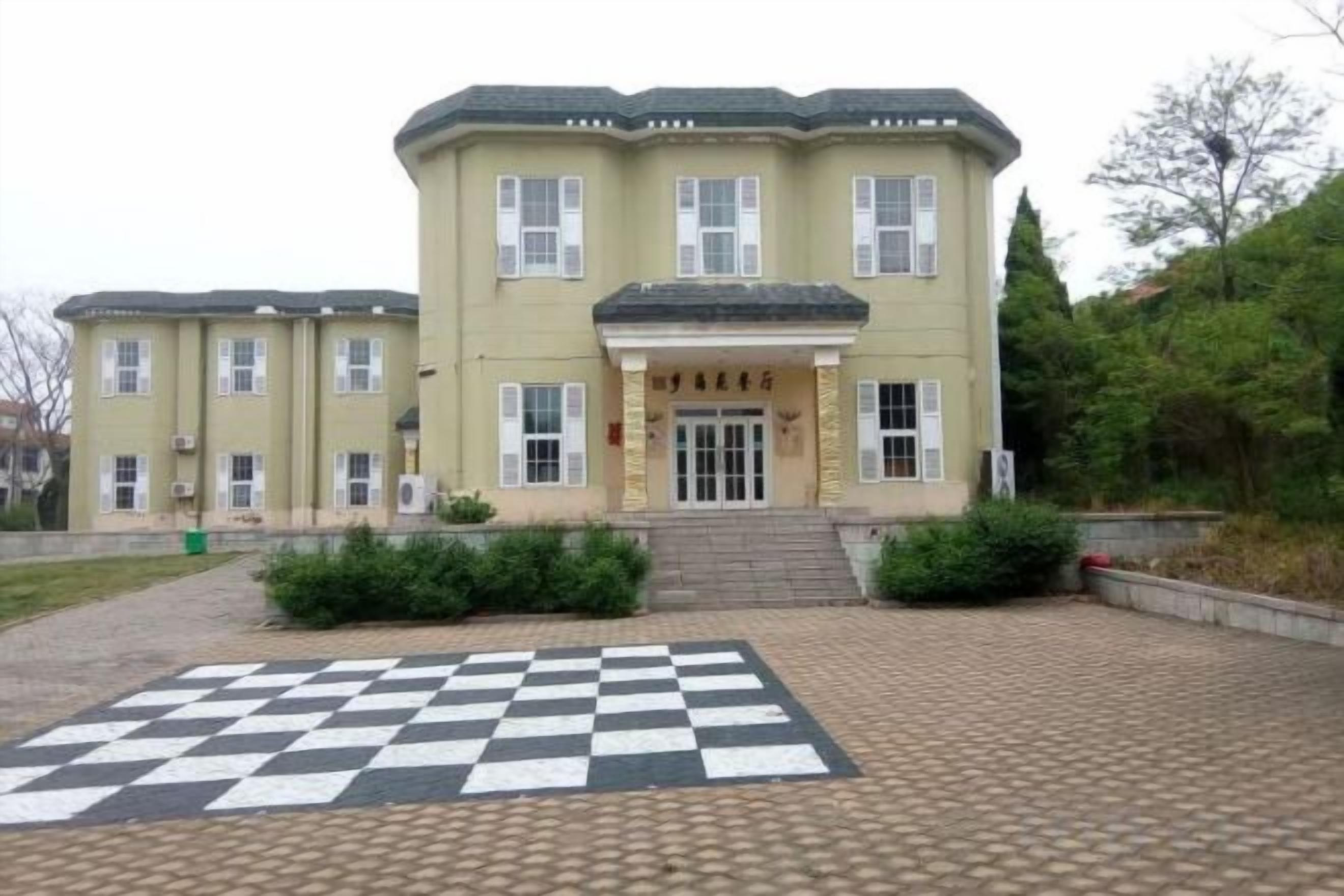 A two-story building with a checkered pavement in front, a set of stairs leading to the entrance, and a small garden area.
