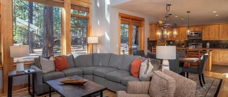 A spacious living area with a large gray sectional sofa, a patterned armchair, and a coffee table. There is a dining area with a wooden table and chairs, and a kitchen with wooden cabinets and a hanging light fixture.