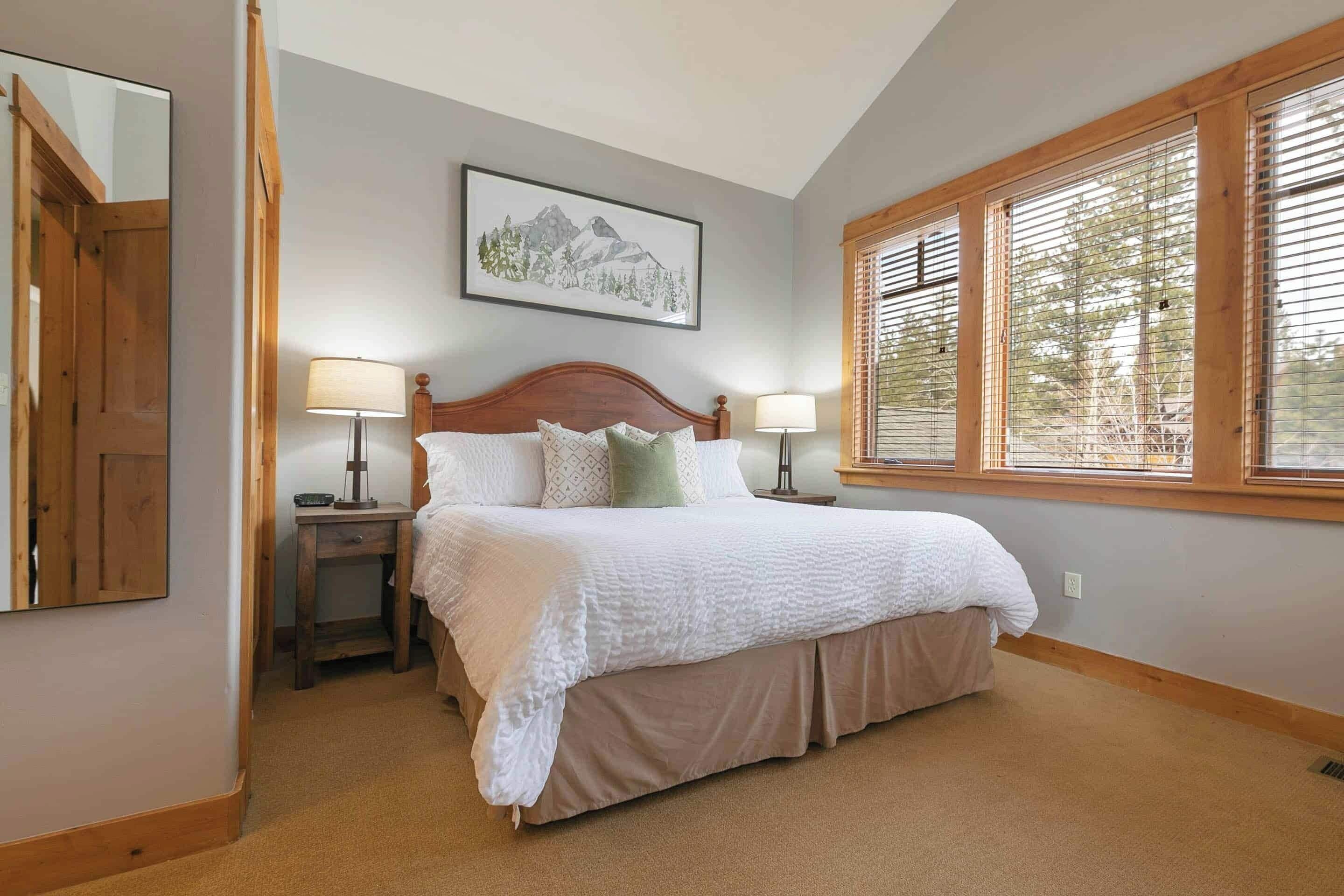 A bedroom with a large bed, two bedside tables, a nightstand, a window with blinds, and a framed picture on the wall.