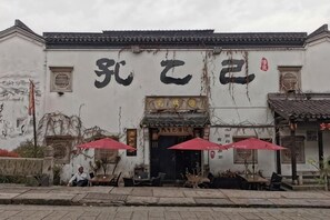 Ein traditionelles Gebäude mit Ziegeldach und einem Schild mit großen Schriftzügen. Es gibt Außenbereiche mit Sitzgelegenheiten und roten Sonnenschirmen.