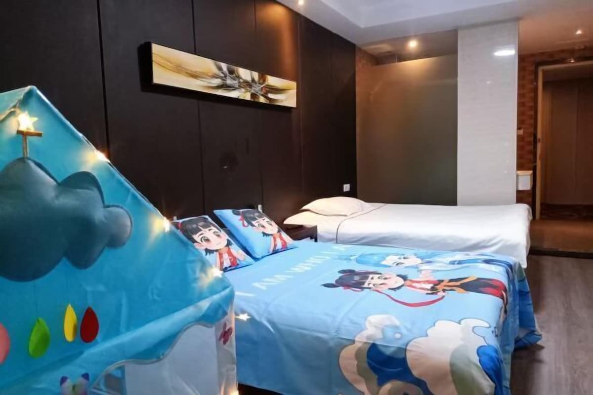 A hotel room with a bed featuring cartoon character bedding, a wall-mounted artwork, and a bedside table with a lamp.