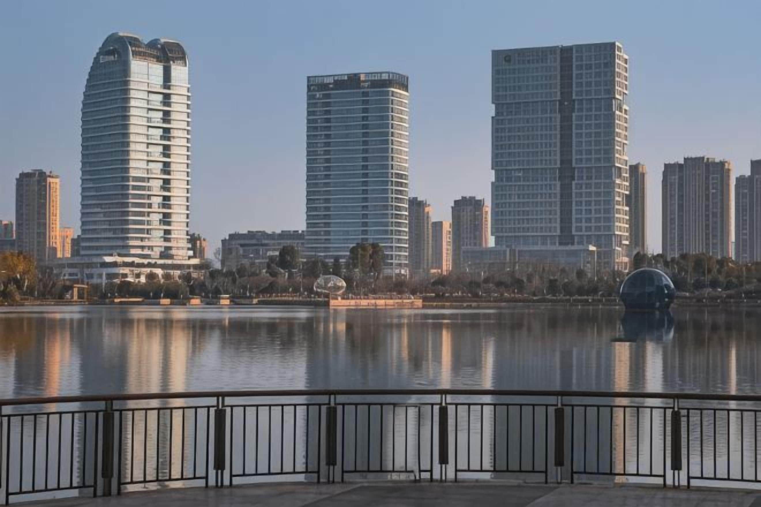 Un paisaje urbano con modernos edificios de gran altura, una masa de agua tranquila y una barandilla de metal en primer plano.