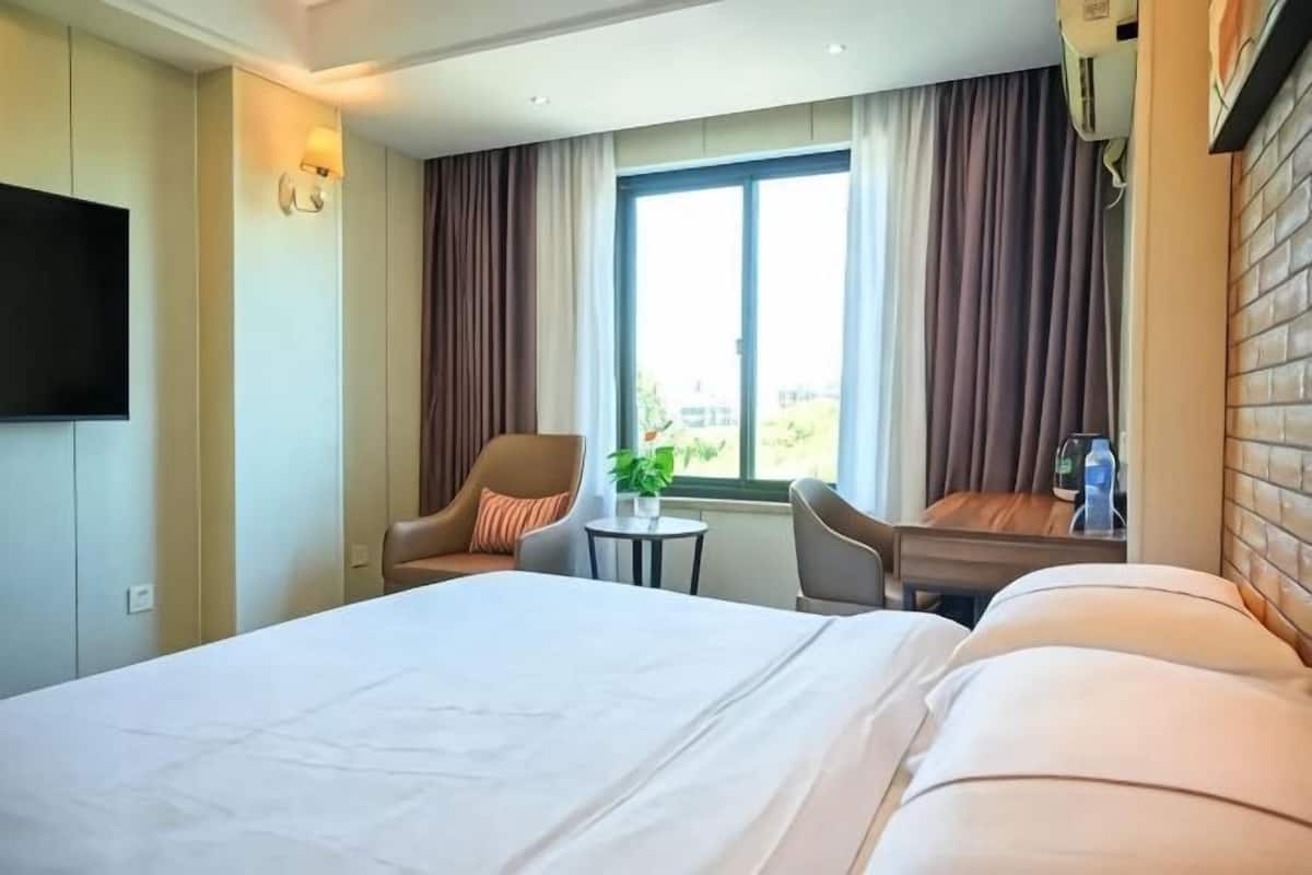 Quarto de hotel com uma cama grande, televisão na parede, uma mesinha com uma planta e uma janela com cortinas.