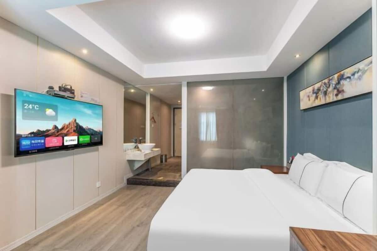 Quarto de hotel moderno com uma cama grande, uma TV montada exibindo a previsão do tempo, um banheiro com pia e espelho, e um quadro na parede.