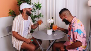 Zwei Personen sitzen an einem runden Tisch mit Getränken, umgeben von Pflanzen und einem weißen Vorhang.