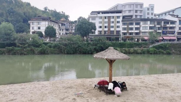 Una playa con una sombrilla de paja, una silla y un bolso en la arena. Al fondo, edificios de varios pisos y balcones.