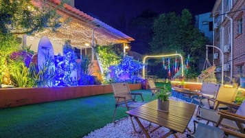 Un patio al aire libre con una mesa y sillas de comedor, rodeado de plantas e iluminado por luces decorativas.