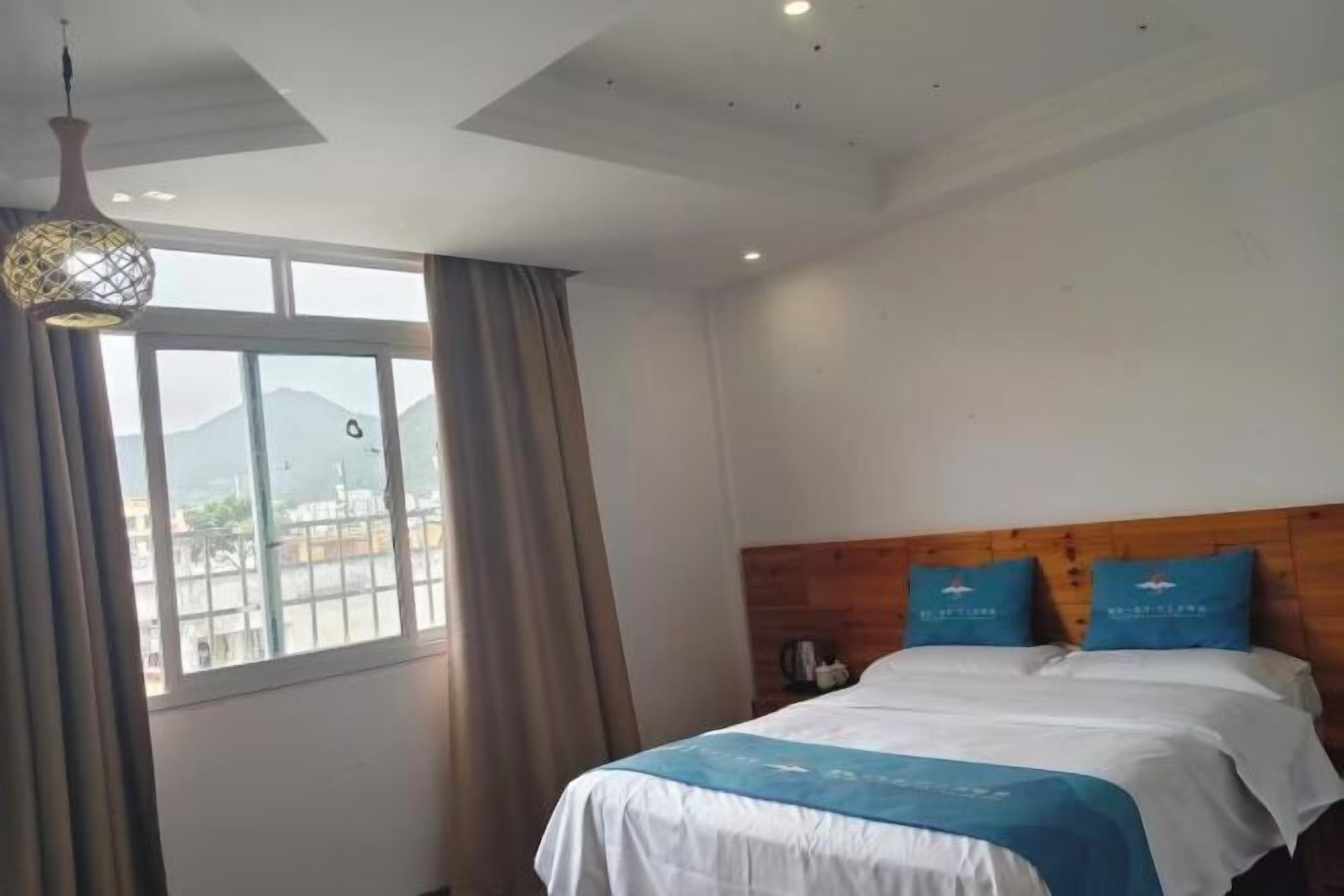 A hotel room with a bed, a wooden headboard, a window with curtains, and a hanging light fixture.