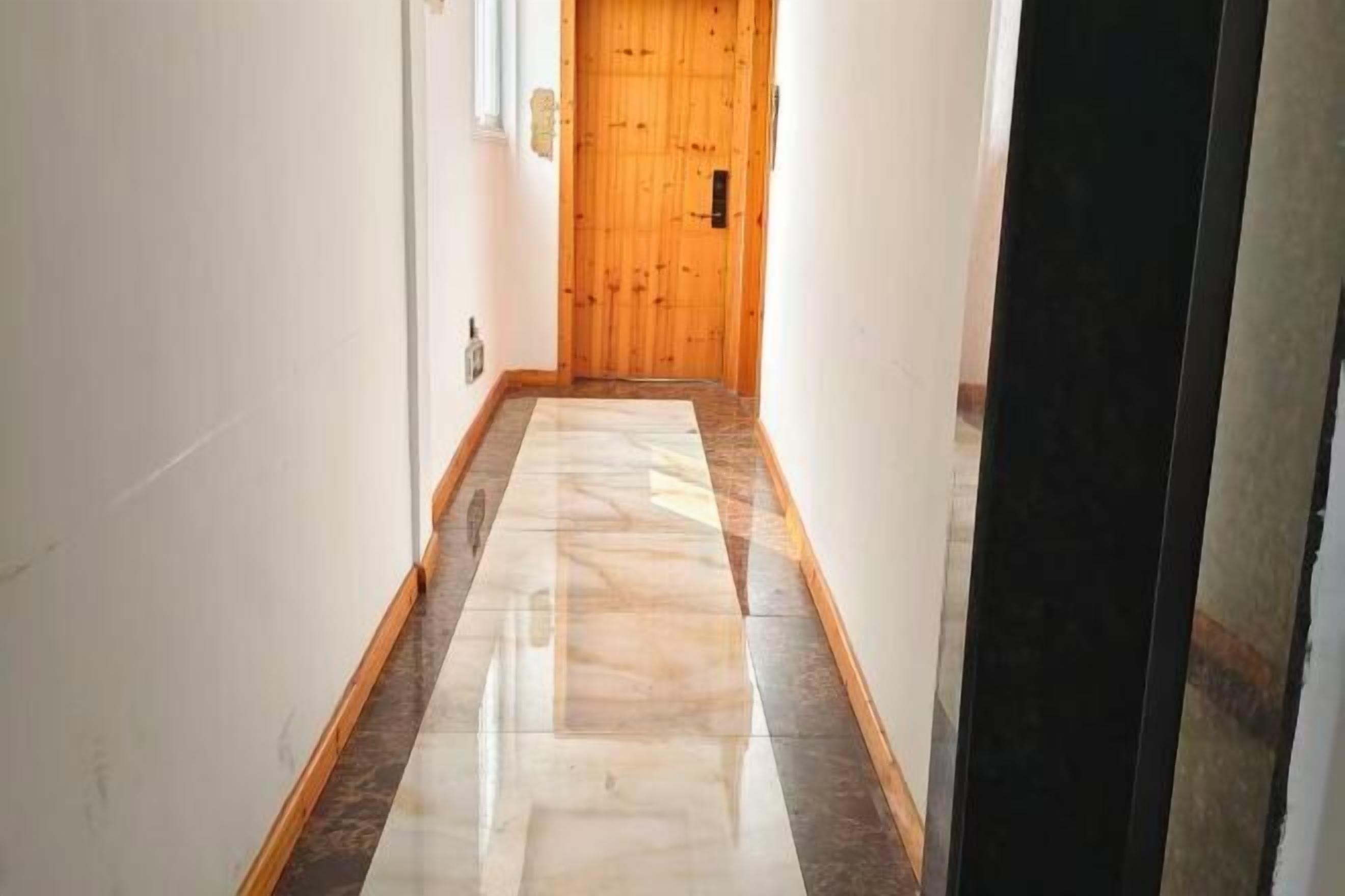 Un corridoio con una porta in legno a fondo, pavimento piastrellato e un interruttore sulla parete.