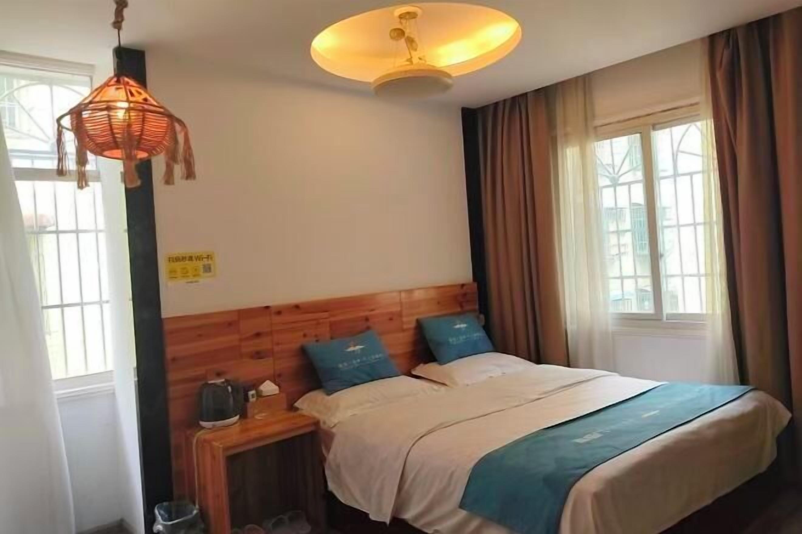 Une chambre d’hôtel avec une tête de lit en bois, un lit avec une literie bleue et blanche, un luminaire suspendu, une table de chevet avec une bouilloire et une fenêtre avec des rideaux.