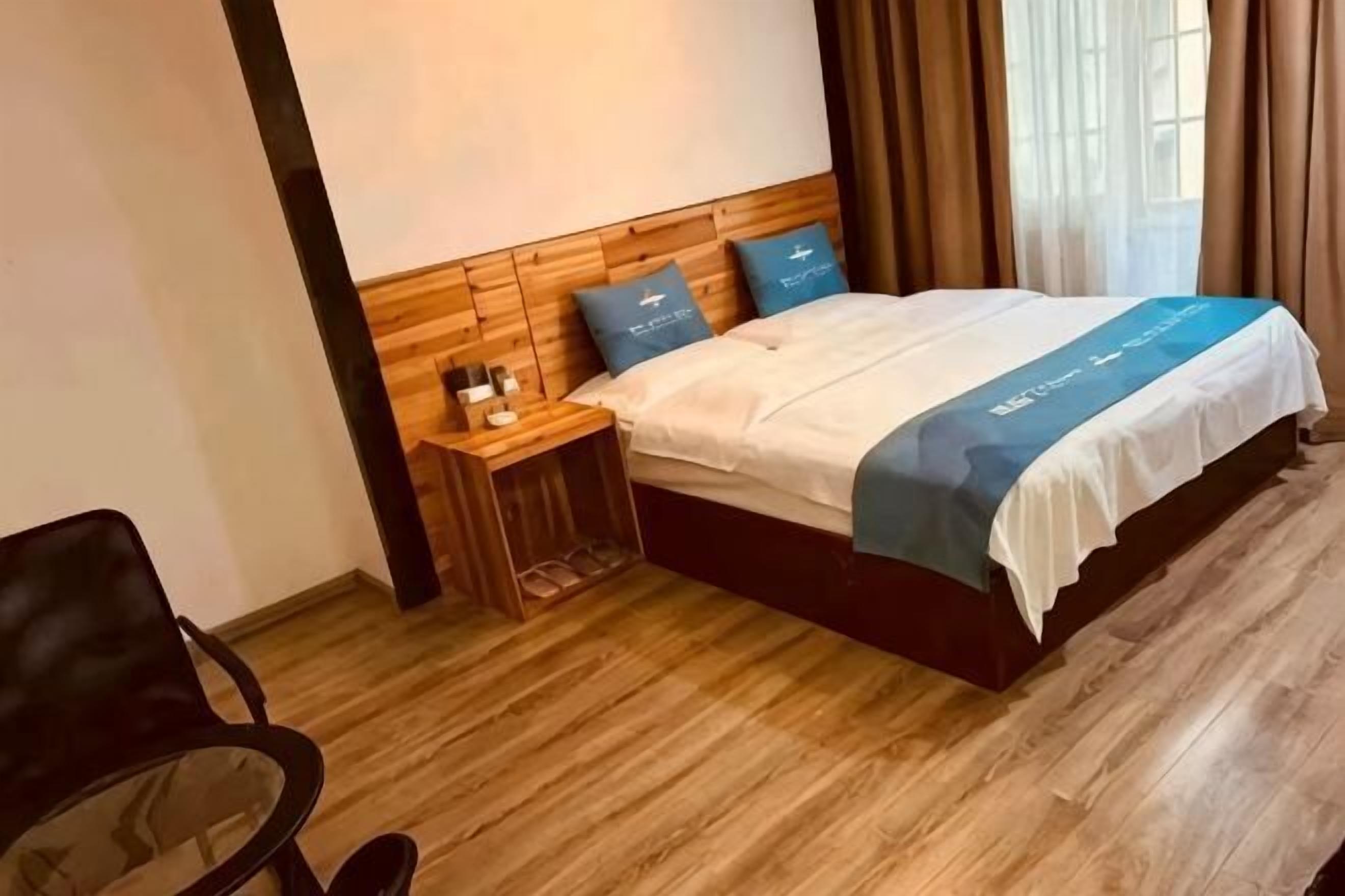 Una habitación de hotel con cabecera de madera, cama con colcha azul y blanca, mesita de noche con lámpara y una silla.