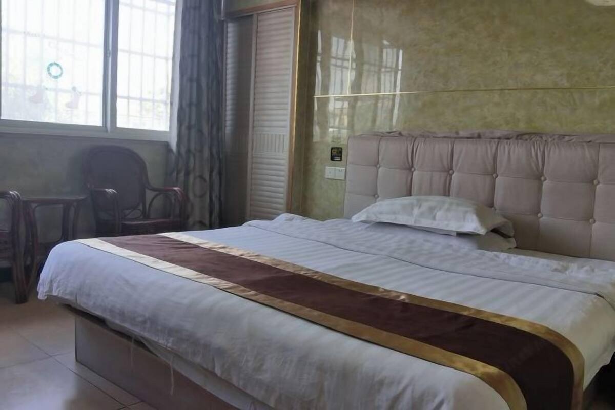 A hotel room with a bed, a chair, a window, and a wall-mounted light switch.