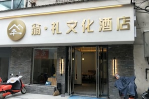 A hotel entrance with a sign above the door, a scooter parked outside, and a person in a blue garment near the entrance.