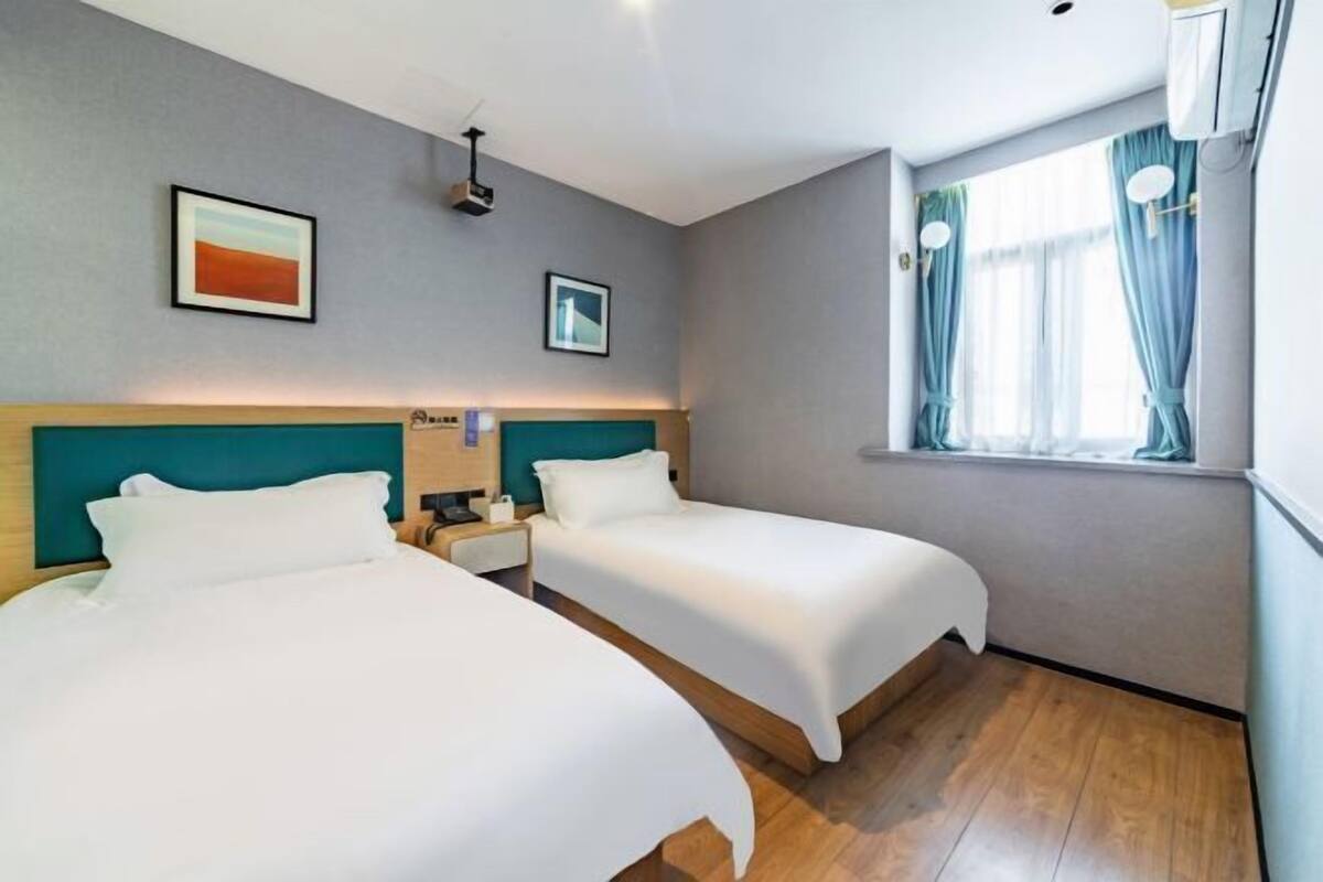 Een hotelkamer met twee bedden, een raam met gordijnen, een schilderij aan de muur en een projector in het plafond.
