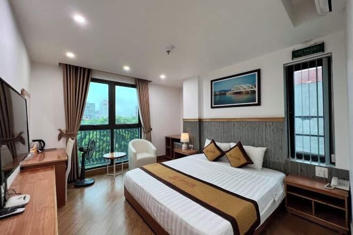 A hotel room with a bed, a desk, a chair, a small table, a lamp, a picture on the wall, and a window with curtains.