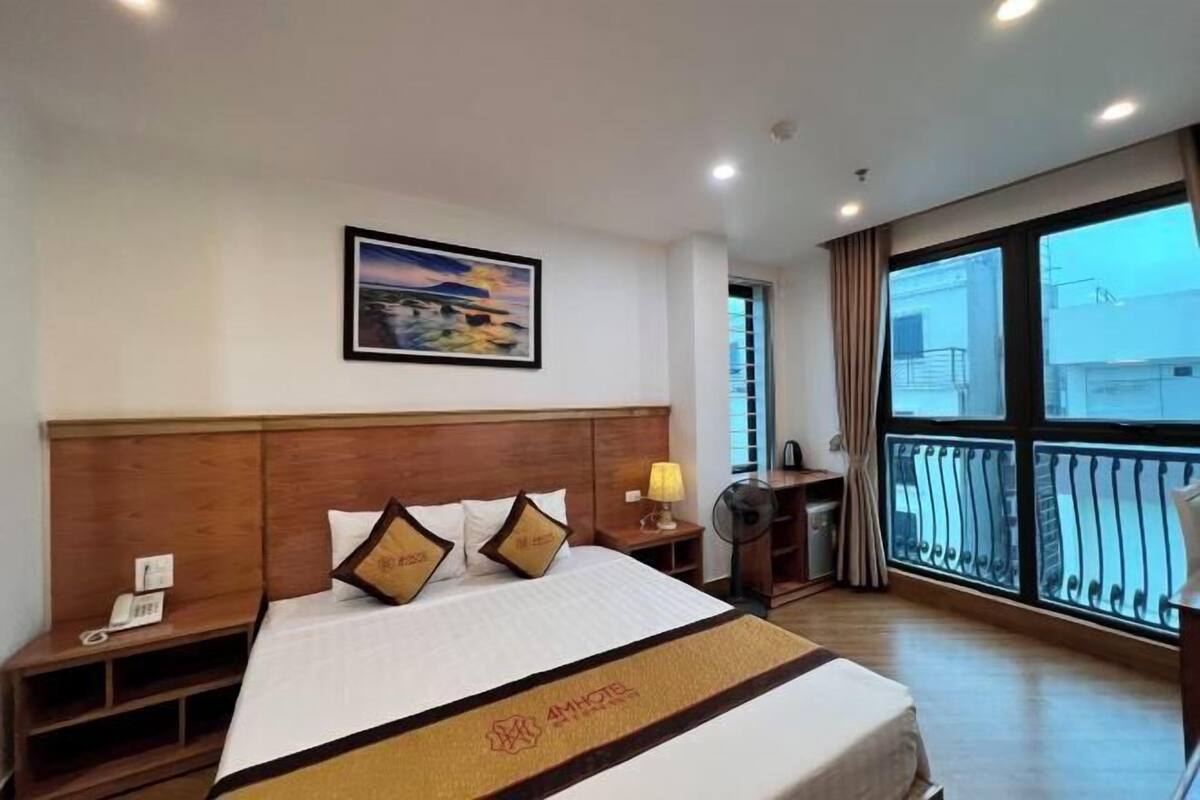 A hotel room with a large bed, a nightstand, a bedside table with a lamp, a fan, and a window with a balcony.