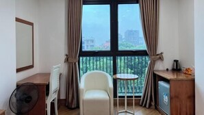 A small room with a large window, a chair, a table, a fan, and a small kitchenette. - Kezan Hotel 4m2 (Vo Cuong)