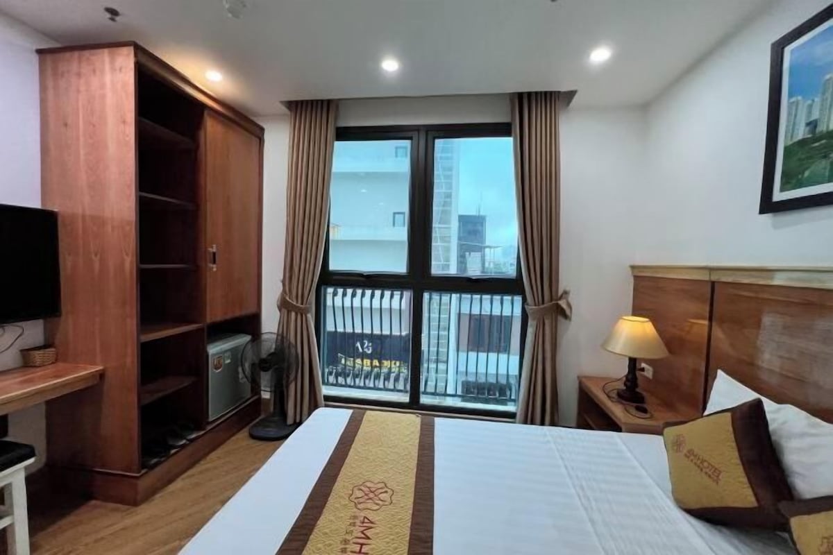 A hotel room with a bed, a wooden wardrobe, a desk with a chair, a TV, a window with curtains, and a nightstand with a lamp.