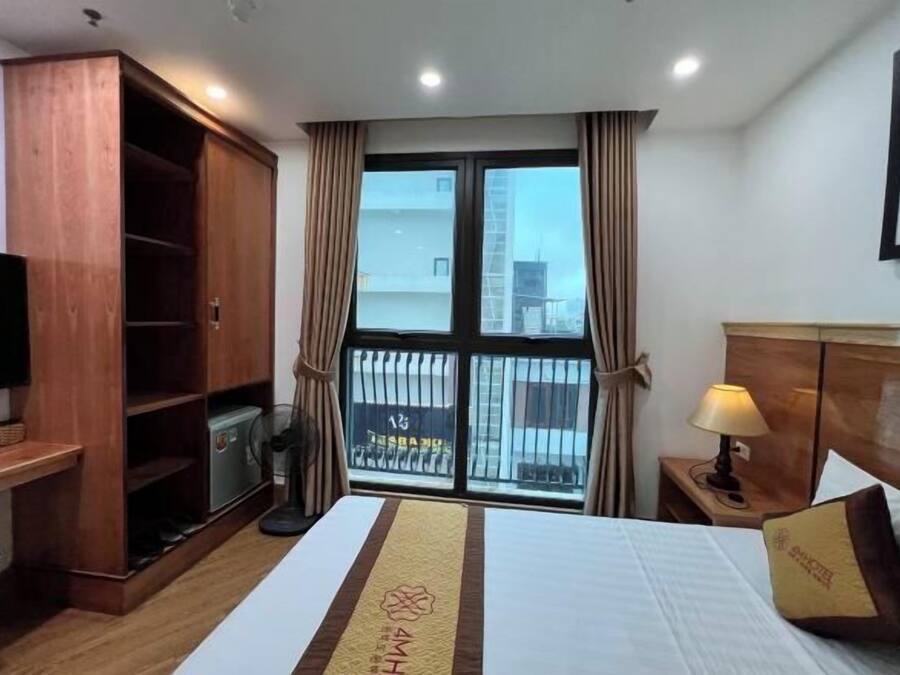 Habitación de hotel con cama, armario de madera, escritorio con silla, televisor, ventana con cortinas y mesita de noche con lámpara.