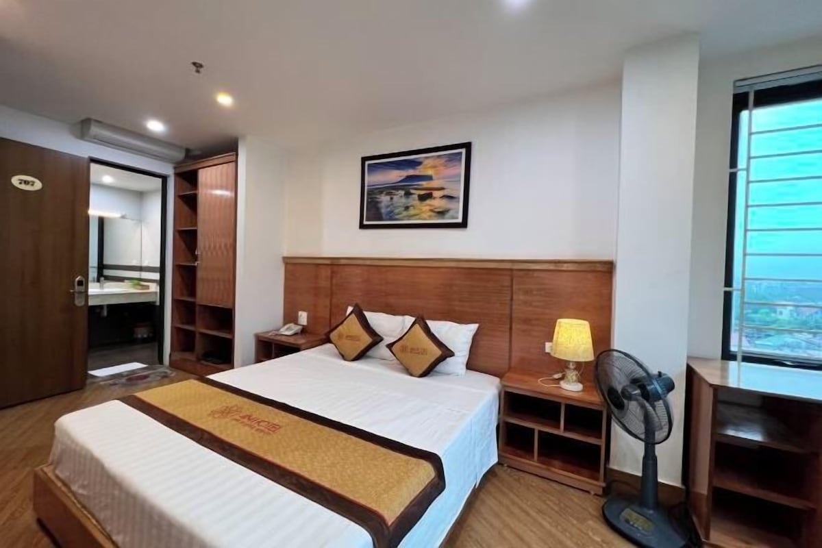 A hotel room with a bed, bedside table, lamp, fan, and a painting on the wall.