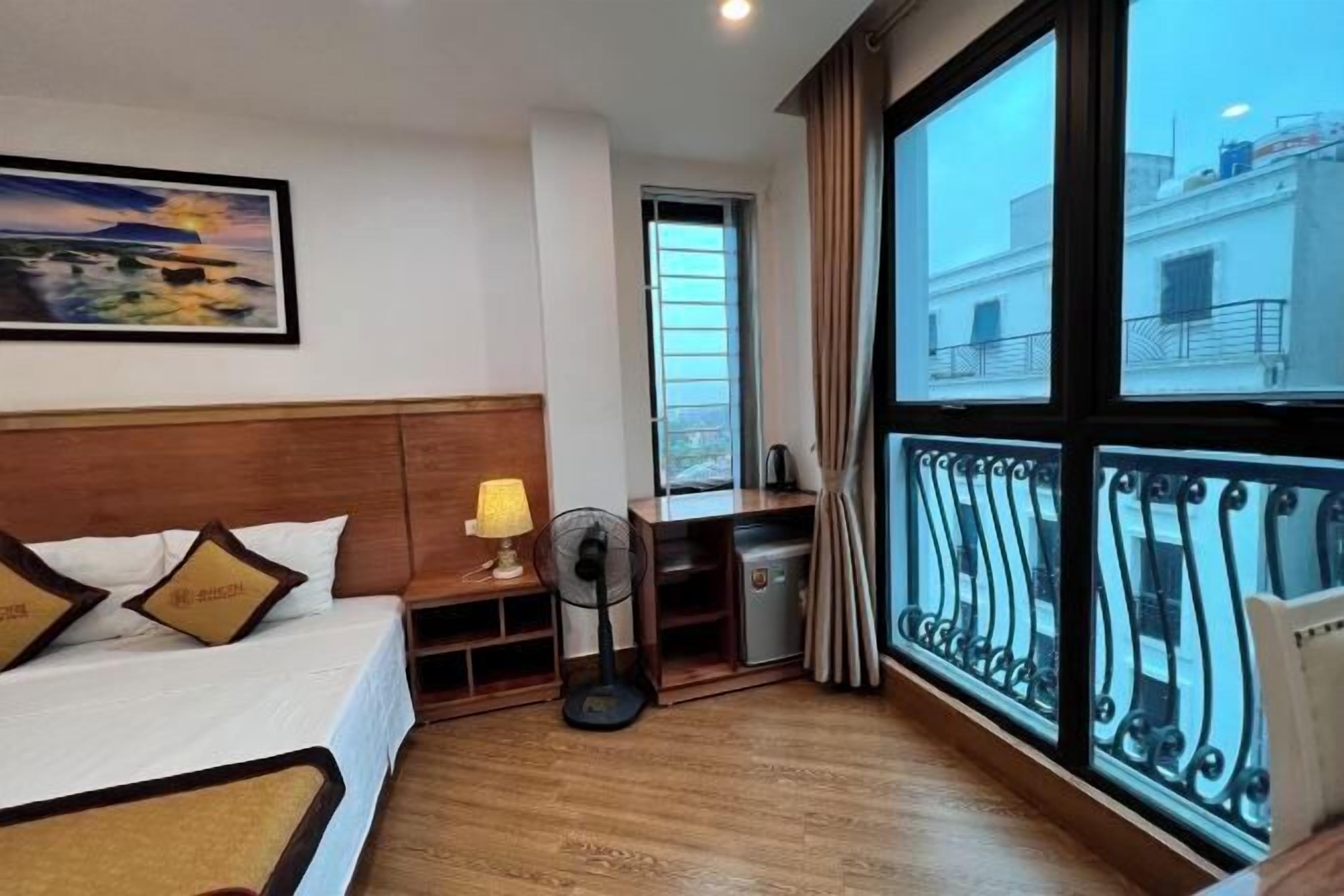 A hotel room with a bed, a desk, a fan, and a large window with a balcony.