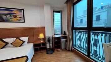 A hotel room with a bed, a desk, a fan, and a large window with a balcony.