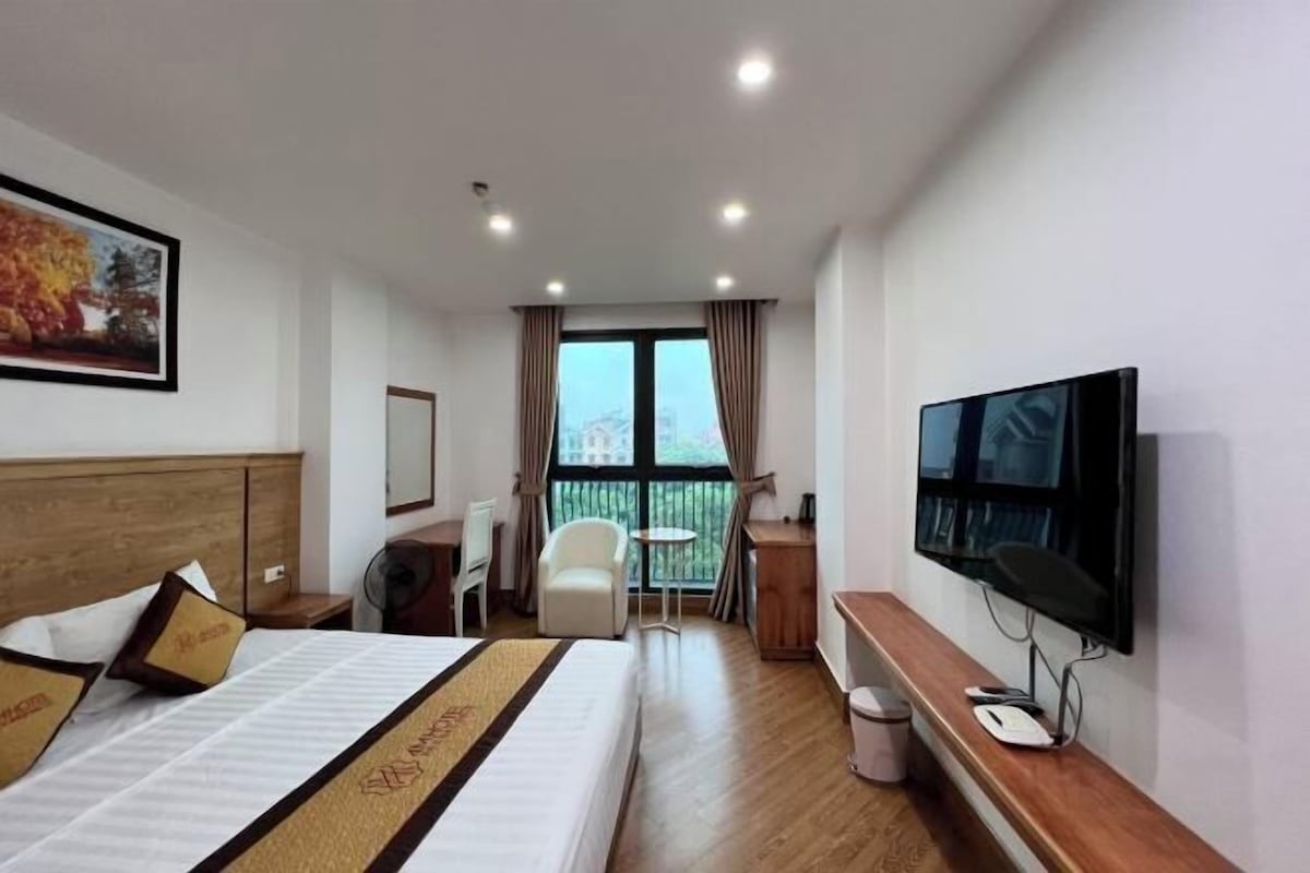 A hotel room with a bed, a TV, a desk, a chair, and a balcony with curtains.