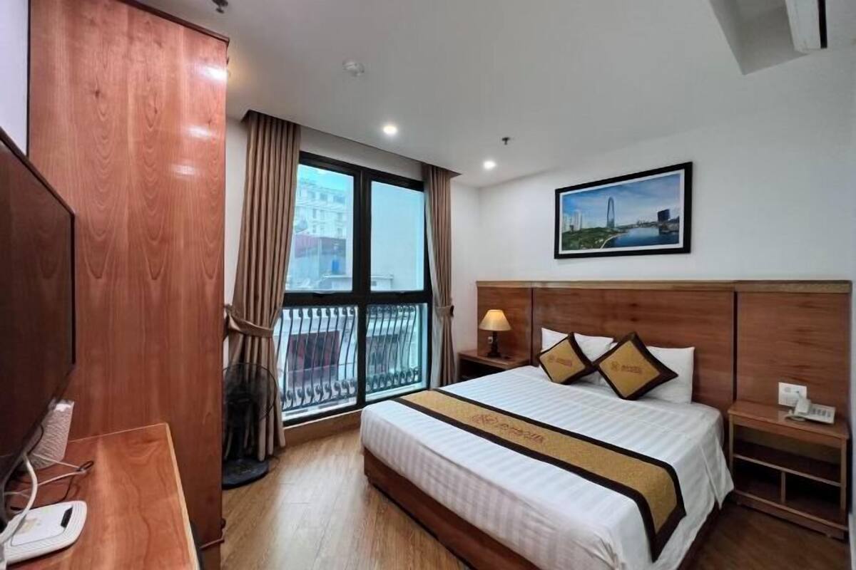 A hotel room with a large bed, a wooden wardrobe, a desk with a telephone, and a window with a view of buildings.